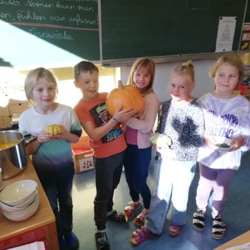 Fünf Kinder stehen vor der Tafel mit Kürbissen in den Händen, neben ihnen ein Topf Kürbissuppe, Teller und Löffel