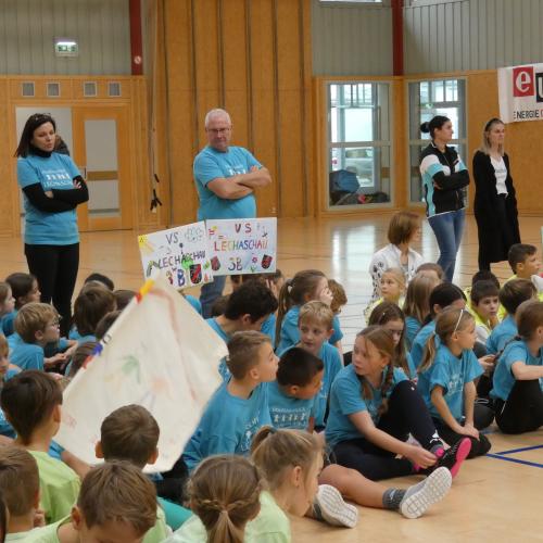 Kinder sitzen in Schul T-Shirts mit ihren Transparenten am Turnsaalboden und warten auf den Beginn