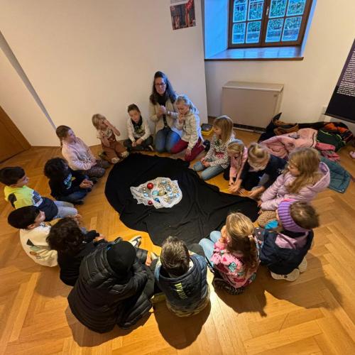Alle Kinder sitzen mit der Museumspädagogin im Kreis am Boden, in der Mitte ein schwarzes Tuch mit vielen verschieden großen bunten Kugeln