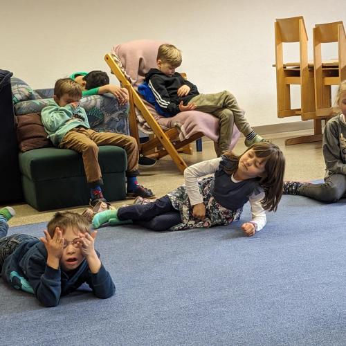 Kinder liegen oder sitzen im Bewegungsraum mit geschlossenen Augen und warten was sie von der Lehrerin bekommen