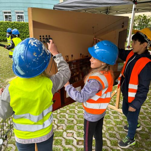 3 Kinder in Schutzausrüstung mit Helm und Warnweste bei einer Schalttafel  - Stromkreise