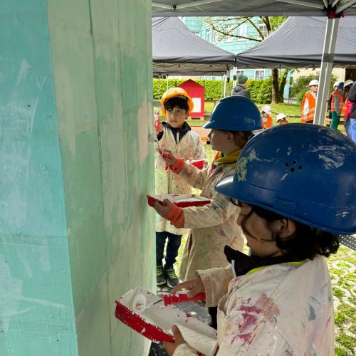 2 Kinder mit blauen Helmen und Malerkitteln beim Streichen einer Wand
