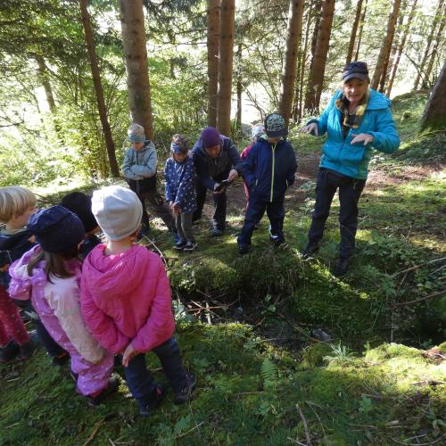 DIe Kinder der Klasse stehen im Wald vor einem Loch, in dem sich eine Quelle befindet
