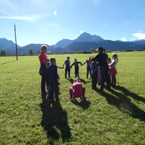 Kindergruppe auf einer Herbstwiese  vor strahlend blauem Himmel mit Bergkulisse. Sie spielen ein Maulwurfspiel