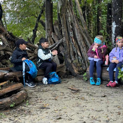 4 Kinder sitzen im Auwald auf Baumstämmen und essen ihre Jaise