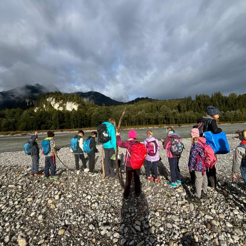 Kindergruppe steht auf einer Schotterbank am Lech, im Hintergrund bewaldete Hügel und toller Wolkenhimmel