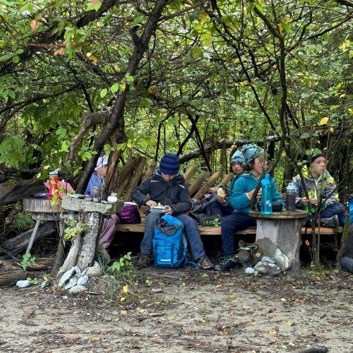 Einige Kinder sitzen auf selbstgebauten Bänken im Auwald und essen ihre mitgebrachte Jause
