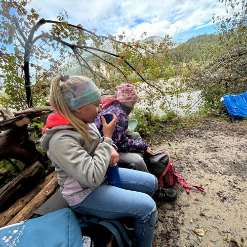 Zwei Kinder sitzen am Lechufer auf Baumstämmen und essen ihre Jause bzw. trinken Tee