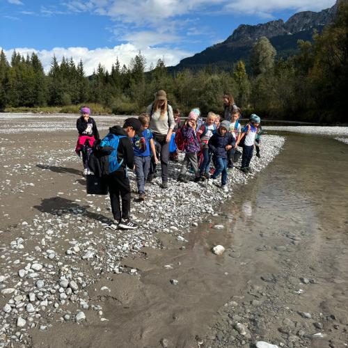 lle Kinder der Klasse sehen an einem Arm des Lech mit zwei Naturpädagogen, im Hintergrund Wald und blauer Himmel mit wenigen Wolken