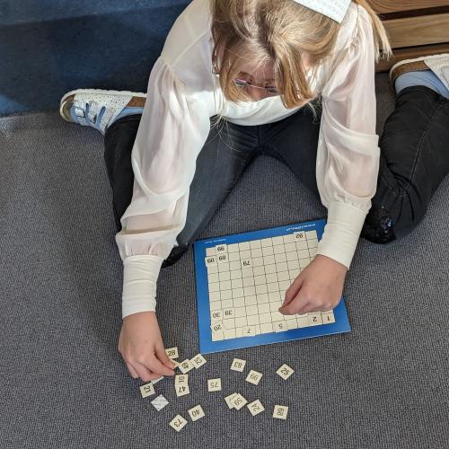 Ein blondes Mädchensitzt am Teppich und legt Zahlenkärtchen auf das Hunderterfeld