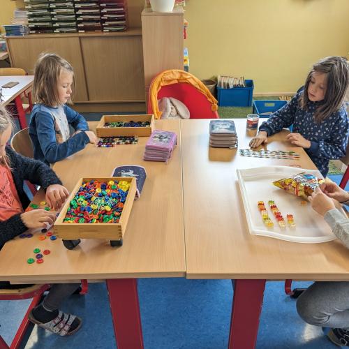 4 Kinder sitzen an einem TIsch  - ein Kind legt 100 mit bunten Knöpfen, ein anderes mit Maiskörnern, ein drittes mit GUmmibärchen und das Vierte Kind mit bunten Büroklammern