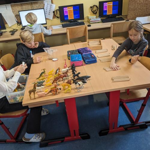 3 Kinder sitzen bei einem Tisch und legen 100 mit Plastiktieren, mit Holzstäbchen und mit Maiskörnern