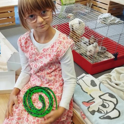 Ein Mädchen sitzt mit ihrer grünen Gummischlange auf der Sch0ß auf der Bank, im Hintergrund ein Käfig mit Henne und 4 Küken