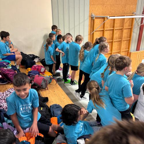 SchülerInnen in baleun Schul T-shirts auf der Tribüne der Mittelschule Königsweg in Reutte, einige stehen am Geländer und schauen den Spilen unten zu, andere jausnen.