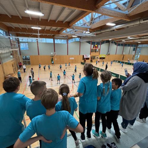 7 Kinder der VS Lechaschau stehen in den blauen T-Shirts der Schule auf der Tribüne der Sporthalle und feuern ein eMannschaft an