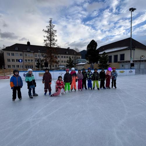 Alle 13 Kinder der Klasse in einer Reihe auf dem Eisplatz, ein Kind sitzt auf einem weißen Schiebehund
