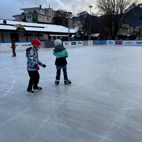 2 Kinder am Eisplatz, im Hintergrund die Bande, Gebäude und ein Baum