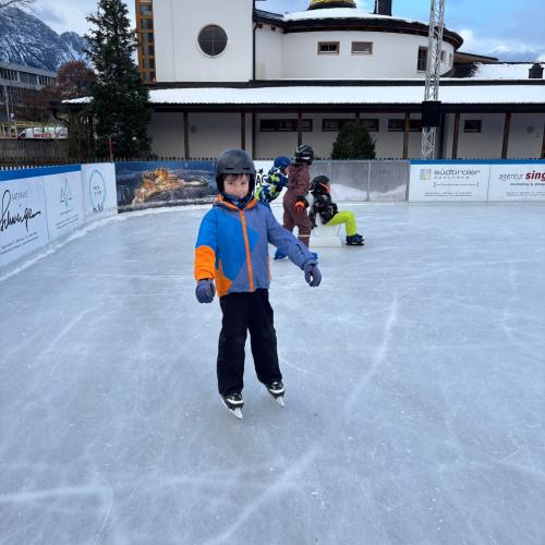 Ein Kind mit Helm, schwarzer Schihose und Blau-orangem Anorak steht am Eis, im Hintergrund weitere Kinder und ein Gebäude