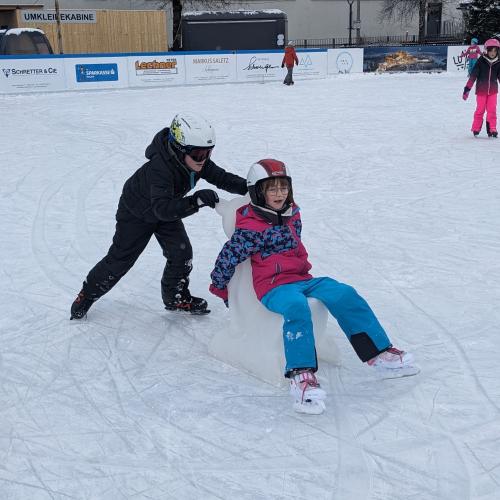 Ein Junge mit Helm und Schibrille schiebt ein Mädchen, das auf einem weißen "Schiebehund" sitzt
