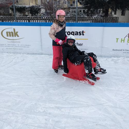 Eislaufülatz - ein Mädchen schiebt einen Jungen, der auf einem roten Schiebehund sitzt