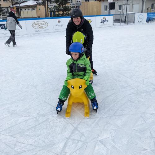 Unsere Schulassistentin schiet gemeinsam mit einem Kind mit gelbem Helm ein en Jungen im grünen Schianzug, der auf einem gelben Schiebebär sitzt