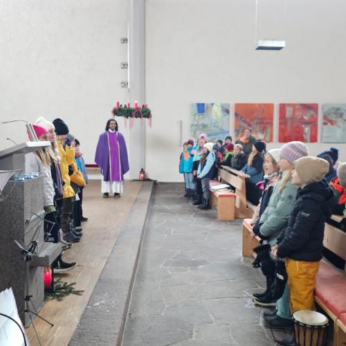 Blick in die Kirche: Der Pfarrer steht hintenin der Mitte uter dem großen hängenden Adventkranz. Links stehen Kinder mit Flöten und singende Kinder, rechts sitzen Kinder in den Bänken und dahinter einige Eltern