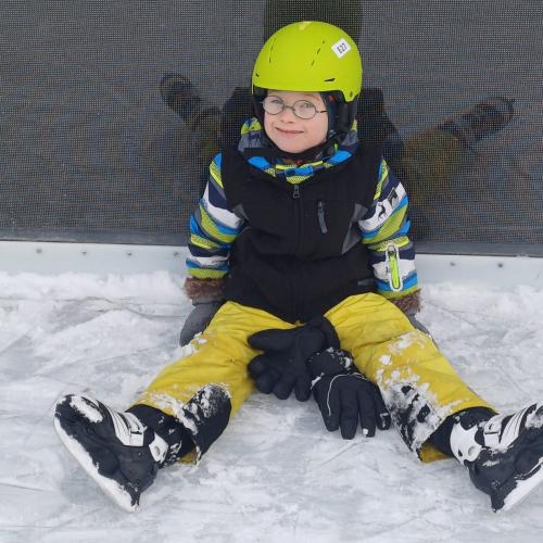 Ein Bub sitzt am Eislaufplatz am Boden - mit einem Grinser im Gesicht - glücklich, aber müde