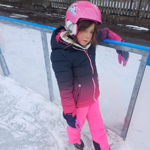 EIn Mädchen in einem rosa Schianzug mit rosa Helm steht mit verlegenem Lächeln an der Bande