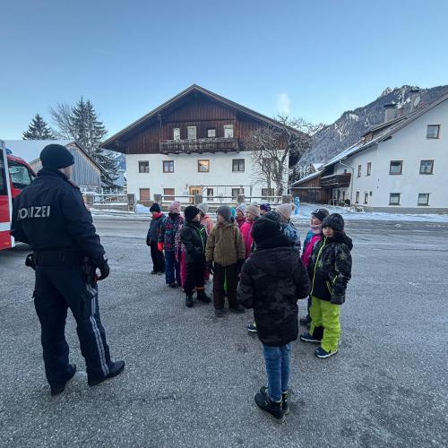 Ein Polizist steht mit den Kindern der Klasse vor der Feuerwehrhalle in Lechaschau, im Hintergrund ein Bauernhaus