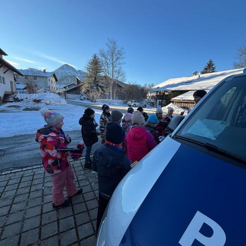 Kinder stehen am Parkplatz neben einem VW Bus der Polizei und hören einem Polizisten zu