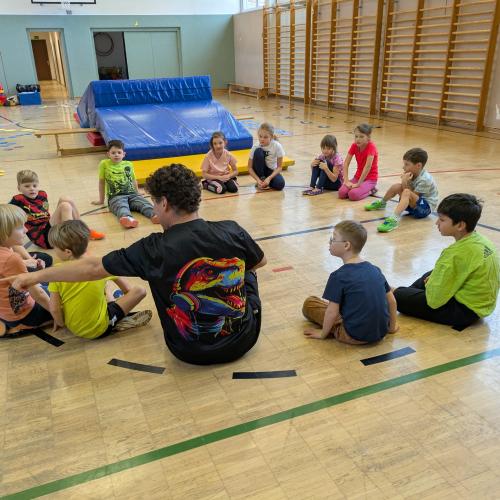 Die Kinder sitzen mit Andi Wagner alias Hopsi Hopper im Kreis am Turnsaalboden und er erklärt das Spiel