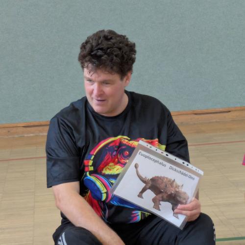 Andreas Wagner "Hopsi Hopper" sitzt am Turnsaalboden mit einem Dinosaureibild in der Hand und erklrt ein Spiel