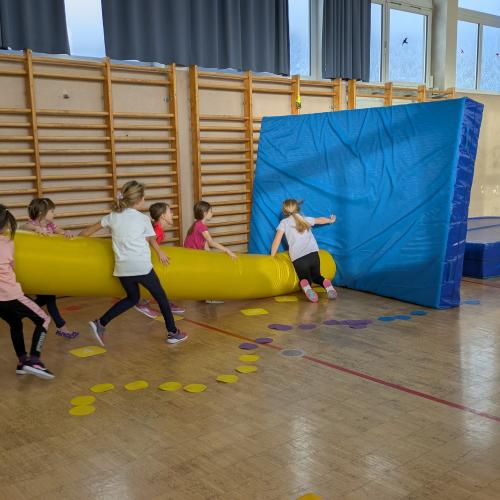 6 Mädchen versuchen im Turnsaal mit einer großen aufgeblasenen Gumminudel eine aufgestelle blaue Matte umzuwerfen