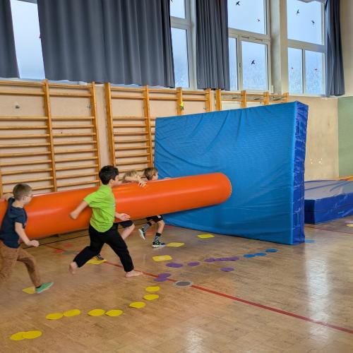 4 Buben versuchen mit einer aufgeblasenen dicken, langen Gummiwurst eine AUfgesellte wieche Matte umzuwerfen