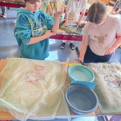 MAn sieht 3 Kinder, die bei ihren Tischen stehen, die mit Folie und Handtuch bedeckt sind un dfilzen mit Wolle