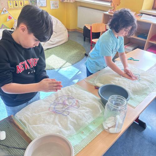2 Kinder stehen bei ihrem TIsch, bedeckt mit Handtuch und Folie sie haben eine Schüssel mit Seifenwasser bei sich und filzen mit Wolle