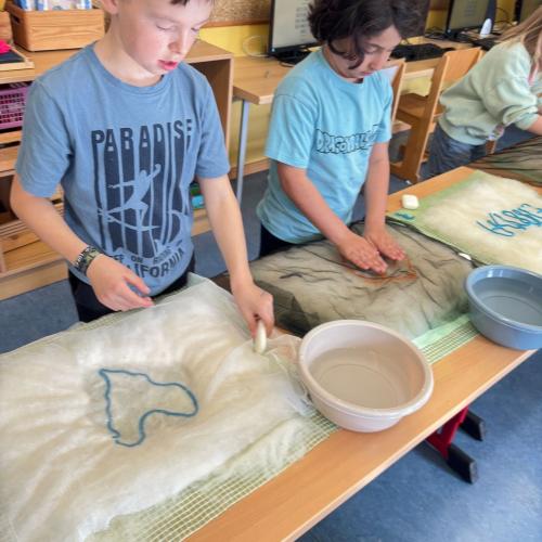 Man sieht zwei Kinder, die bei ihrem Tisch stehen, eine Schüssel mit Seifenlauge am Tisch und das Arbeitsstück. Ein Kind hat ein Herz auf eine Unterlage gefilzt
