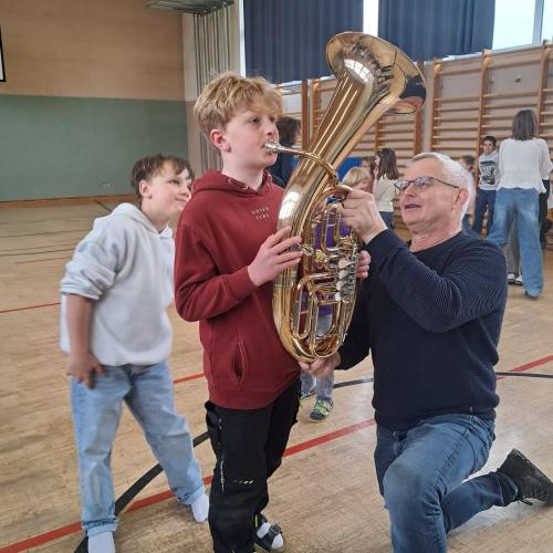 Ein Junge versucht den Kinderbass zu spielen, ein Mitglied der MK hätlt das Instrumen, zwei weitere Kinder warten