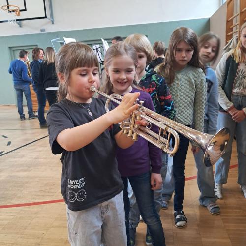 Ein Mädchen bläst mit dick aufgeblasenen Backen das Tenorhorn, eine Schlange Kinder wartet 