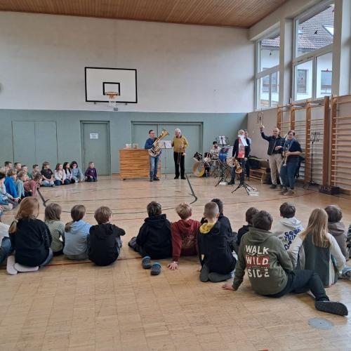 VIele Schüler:innen sitzen am Boden des Turnsaales im Kreis und beim Eingang haben sich die 7 Musikanten mit ihren Instrumenten aufgestellt