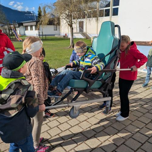 Am Schulhof, ein Junge sitzt im Roll-/Tragestuhl des Rettungswagens, MitschülerInnen tragen ihn.