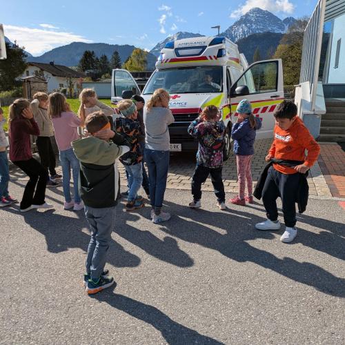 Am Parkplatz vor der Schule steht eij Rettungswagen, die Kinder der 2a Klasse stehen davor und halten sich die Ohren zu, das das Signalhorn eingeschaltet ist.