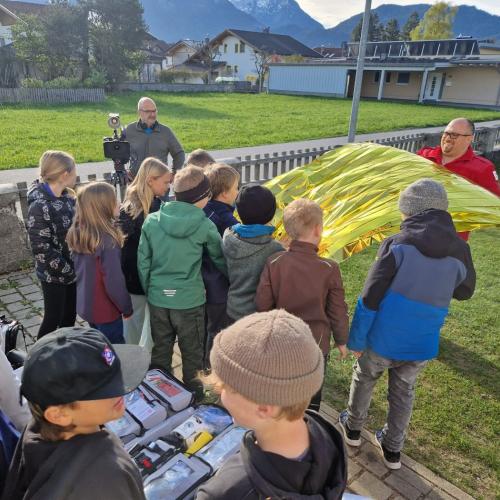 SchülerInnen der 4. Klasse stehen mit einem RotKruemitarbeiter im Schulhof und Betrachten die Rettungsdecke