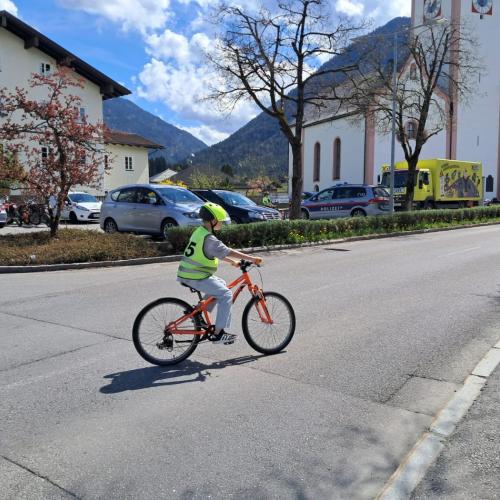 Ein Kind mit gelber Warnweste mit der Nummer 5 fährt gerade vom Parkplatz beim Widum in Breitenwang auf einem orangen Rad. Im Hintergrund die Dekanatskirche BReitenwang und das AUto der Raiffeisen Verkehrsschule