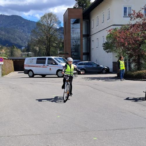 Man sieht zwei Kinder, die auf ihren Fahrrädern fahren, bekleidet mit Warnwesten, fahren, ein Kind gibt ein Handzeichen zum Abbiegen, im Hintergrund das Widum von Breitenwang