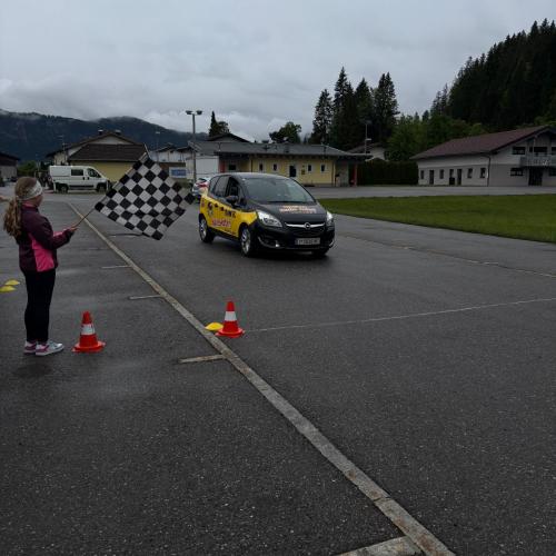 Straße beim Sportplatz, regnerisches Wetter, grauer Himmel, das ÖAMTC AUto fährt, links stehen Kinder mit einer Fahne mit schwarz-weißem Karo