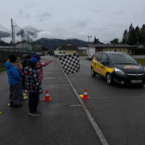 Unfreundliches Regenwetter, Straße beim Sportplatz, das ÖAMTC Auto "Hallo AUto" ist rechts im Bild, links stehen wartende Kinder
