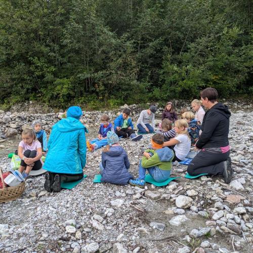 Alle Kinder der Klasse sitzen im Kreis im Schotter beim Bach und besprechen die Untersuchungsergebnisse