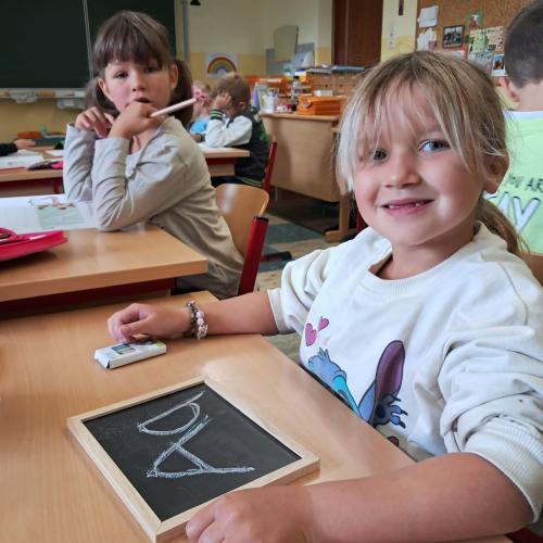 Ein Mädchen schreibt den Buchstaben a auf einer kleinen Tafel, im Hintergrund sitzen zwei weitere Kinder