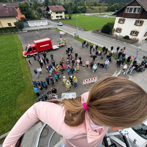 Im Vordergrund sieht man ein Mädchen in der Höhe des zweiten Stockes im Bergekorb. Dahinter - Blick nach unten wo die bereits "evakuierten" Kinder am Parkplatz stehen. Auch ein Feuerwehrauto steht dort.